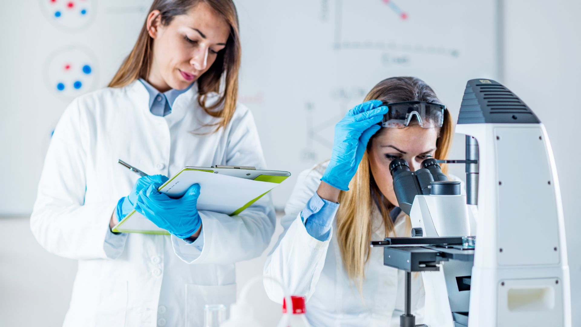 Two women in a laboratory. One looking into a microscope and the other writing on a clipboard