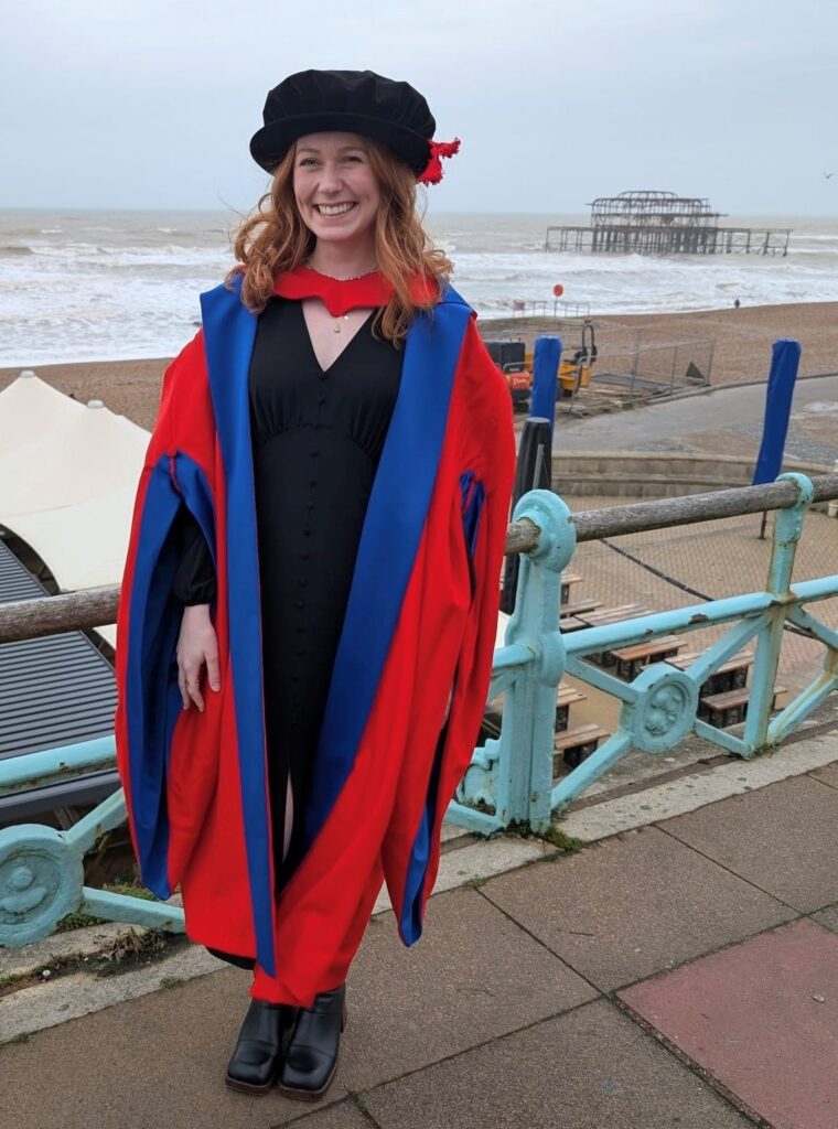 An image of Libby standing on Brighton seafront in her graduation robes as she celebrates completing her PhD.
